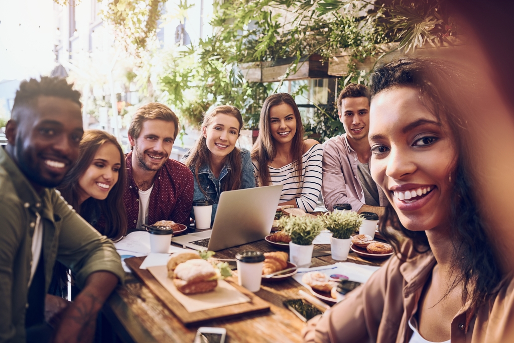 people smiling, sitting around a table with a laptop and food